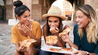 Three beautiful women sitting on the stairs of the city streets eating pizza from a street stall. The happy girls enjoy the week