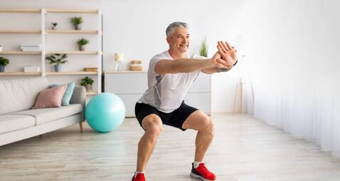 Strength workout concept. Active mature man doing squats at home, exercising in living room, panorama, free space
